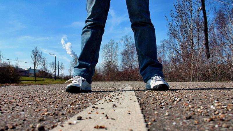 iemand staat met benen gespreid boven een streep op de weg (alleen benen/spijkerbroek en schoenen zichtbaar) op de achtergrond blauwe lucht en kale bosjes en bomen
